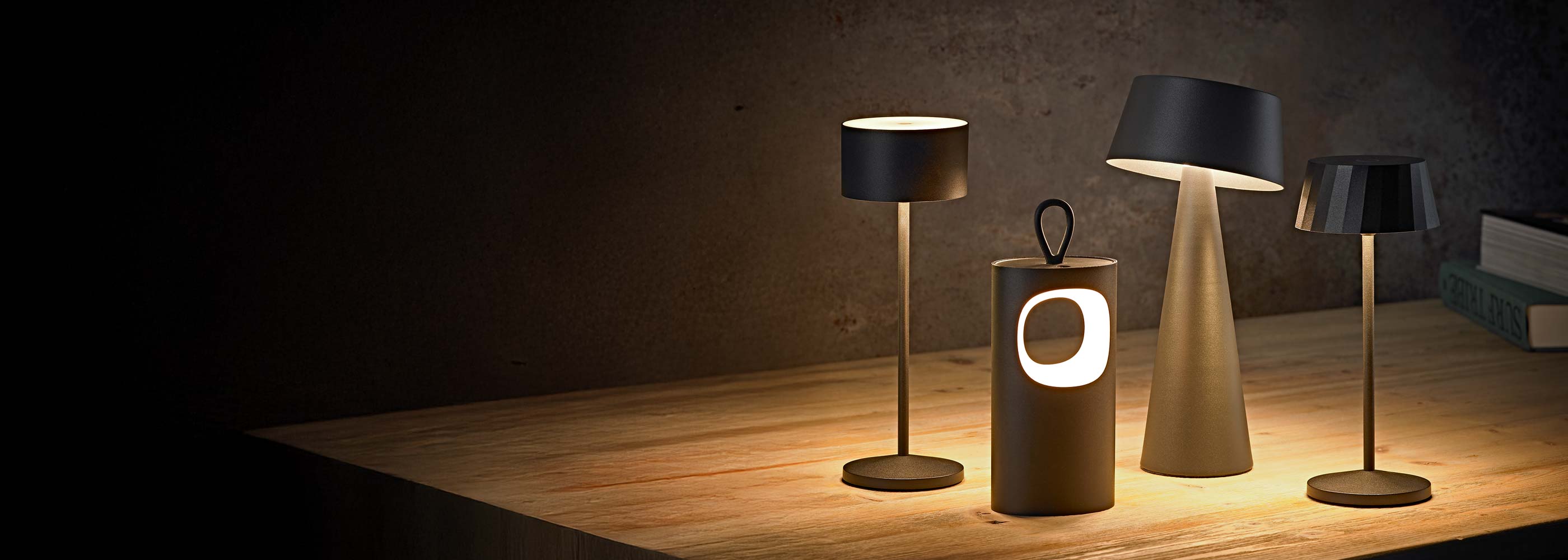 Vier kabellose Akku-Tischleuchten der Sigor Light Collection auf Holztisch vor dunkler Wand: Veluna, Lunari, Solino und Facetta (von links nach rechts), warmweißes Licht schafft gemütliche Atmosphäre