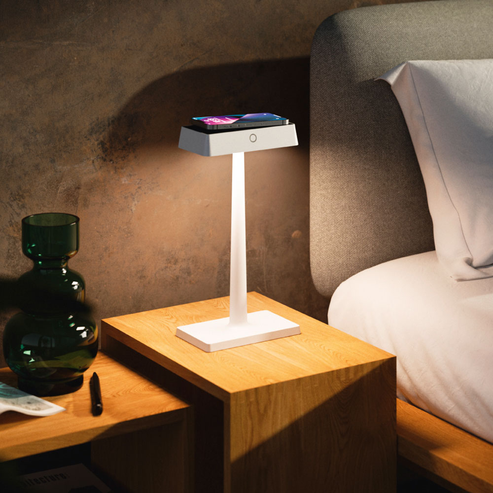 Weiße Sigor Nucharge Akku-Tischleuchte auf einem Nachttisch aus Holz neben dem Bett, auf ihrem Kopf liegt ein Smartphone zum Laden